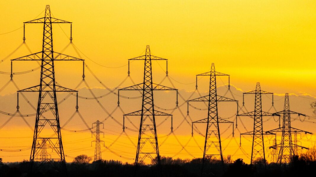 Six electricity pylons are silhouetted at sunset. The pylons are dark against a yellow sky.