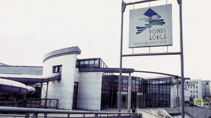 The entrance to Ponds Forge International Sports Centre in Sheffield is glass-fronted with white brick on either side. A purple water slide snakes out of the building and back in again, on the left-hand side of the image.