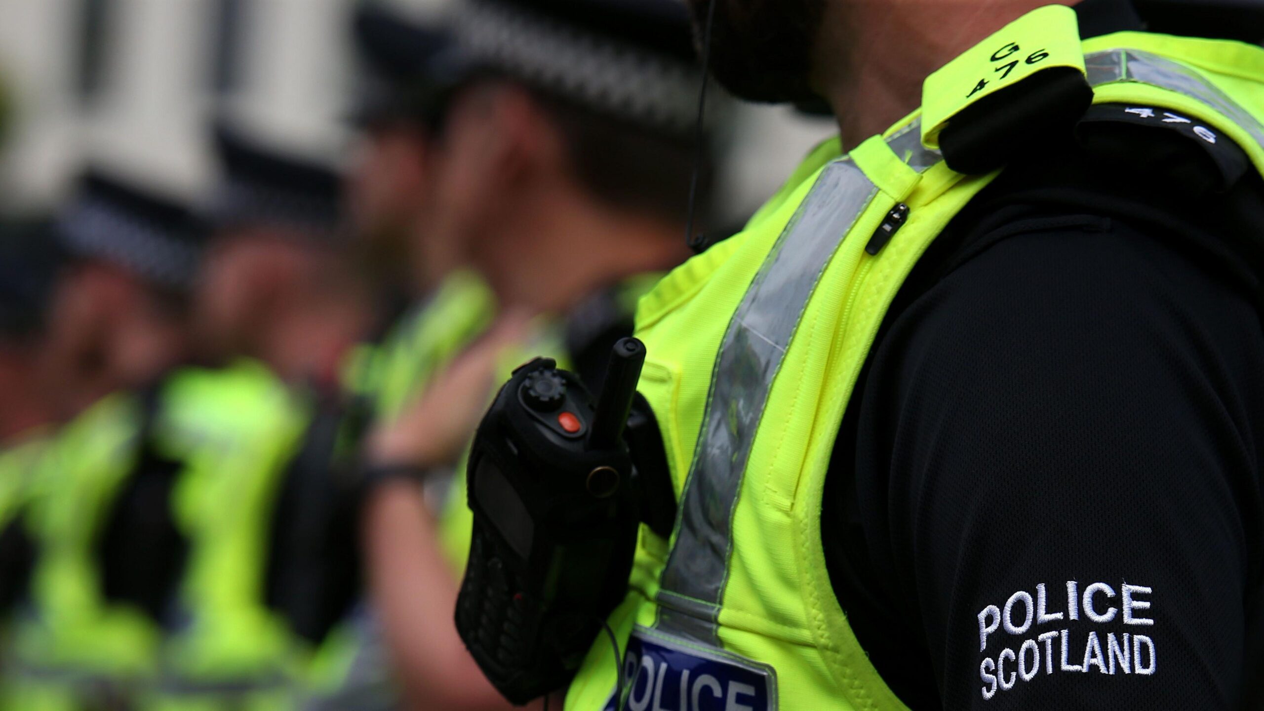 A row of police officers wearing hi-viz vests over black shirts with the words 'Police Scotland' in white text on the arm.