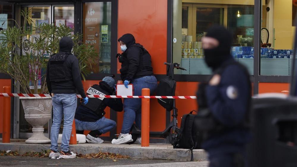 Three police officers crouch down near the wall of a shop. Red and white tape secures the area. Another officer wearing a black balaclava can be seen in the foreground.