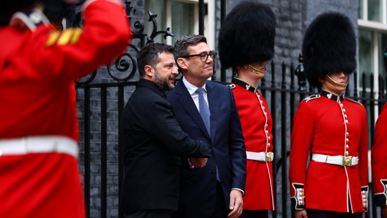 Volodymyr Zelensky and Andy Burnham shaking hands outside the front door of Number 10 Downing Street looking off camera. On either side of both men are soldiers from Number 12 Company Irish Guards Westminster wearing their red uniforms with bearskins.