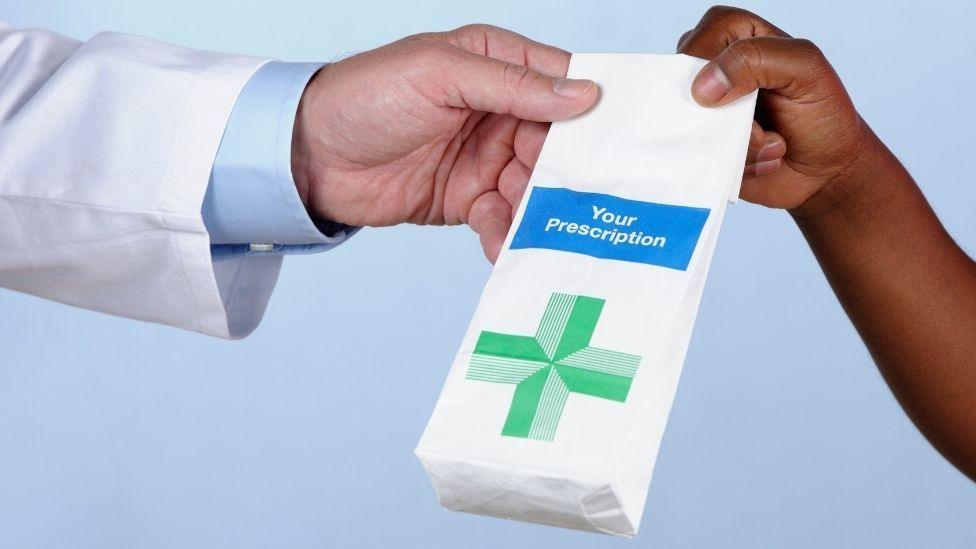 A paper bag with the words "your prescription" written in white within a blue box. There is a green cross symbol on a white paper bag.