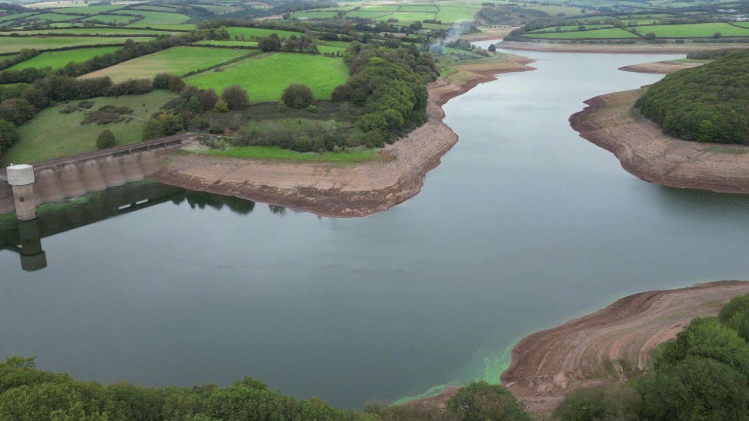 The image shows an aerial view of a reservoir surrounded by patchwork farmland and wooded hills, with large areas of exposed brown shoreline visible around the water's edge. A dam structure can be seen on the left side of the reservoir.