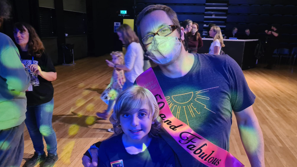 Brown with short brown hair, brown glasses and a covid mask on stands beside Bertie, who has long blond hair. The pair appear to be at a gig with strobe lighting going off around the room.