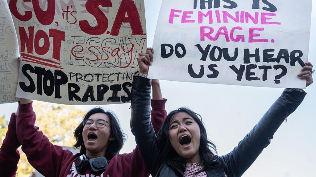 Two protesters hold up signs, with one saying "CU , it's S.A. not essay. Stop protecting rapists" and the other saying "This is feminine rage. Do you hear us yet?"