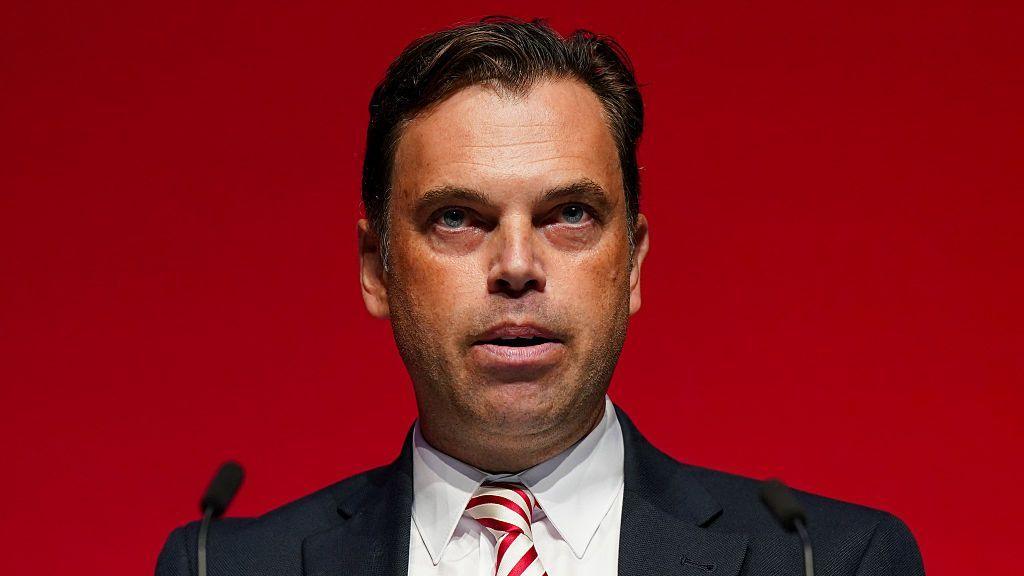 Ken Skates is a man with brown hair, a navy suit, and a red and white-striped tie standing in front of a red background.