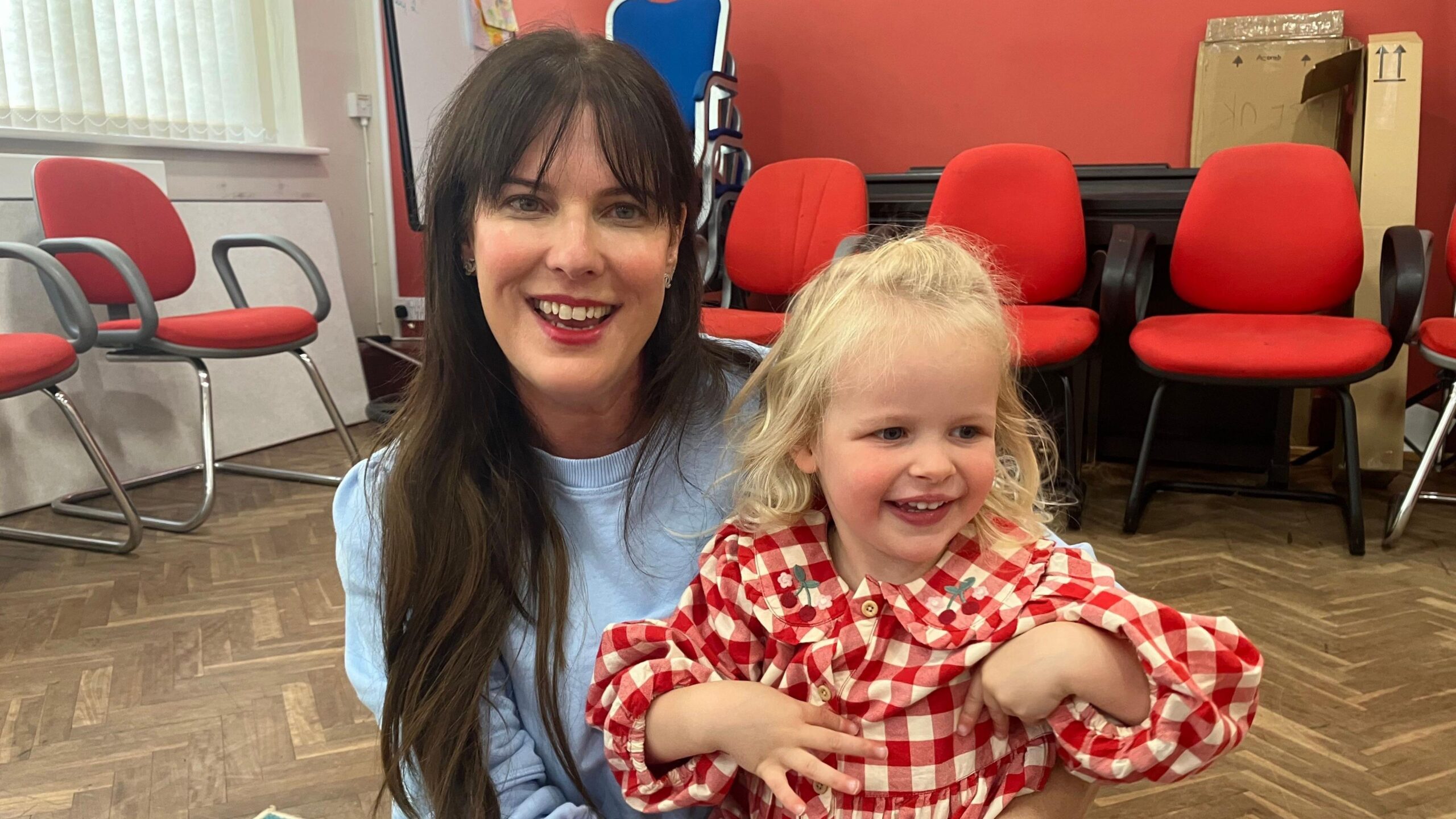 A mum with blue top and long brown hair, sits on the floor of a church hall with her three year old daughter on her knee, who is wearing a red gingham dress and has blonde hair. There are red chairs lining the walls behind her.