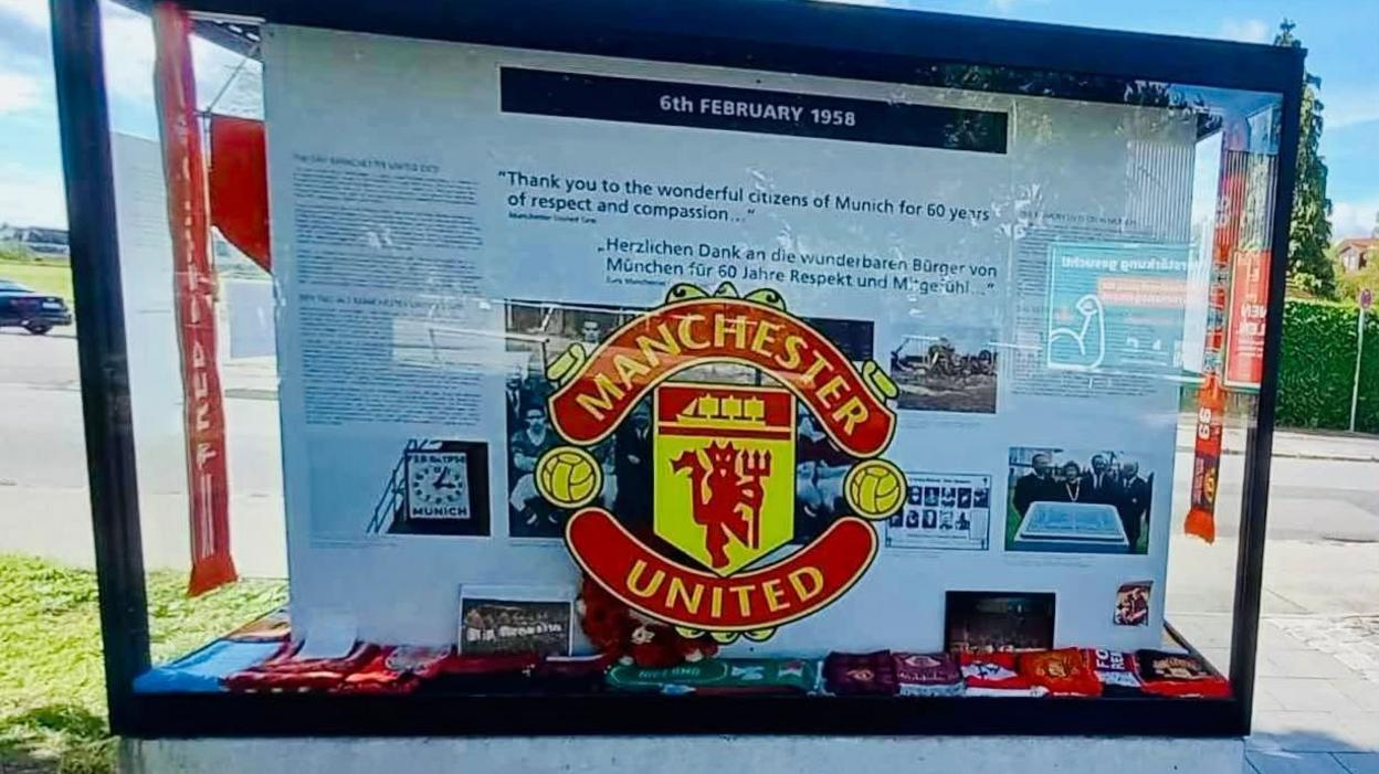 A large rectangular glass display case with a black metal frame stands on a grey concrete base outdoors at Manchesterplatz in Munich. A large red, yellow and black Manchester United crest is displayed prominently in the centre. Above it is a black panel reading "6th February 1958", with historical information, photographs and tributes arranged behind the glass.