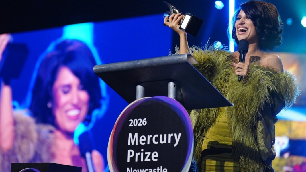 6 Music Festival 2027 heads to Newcastle Raye standing on stage holding a small, silver trophy. She is laughing with a big smile on her face and wearing a green, feathered dress. She has dark, short hair and is holding a microphone. There is a podium in front of her with a circular sign which reads 2026 Mercury Prize Newcastle. There is a large screen to the left which shows Raye with her eyes closed and smiling.