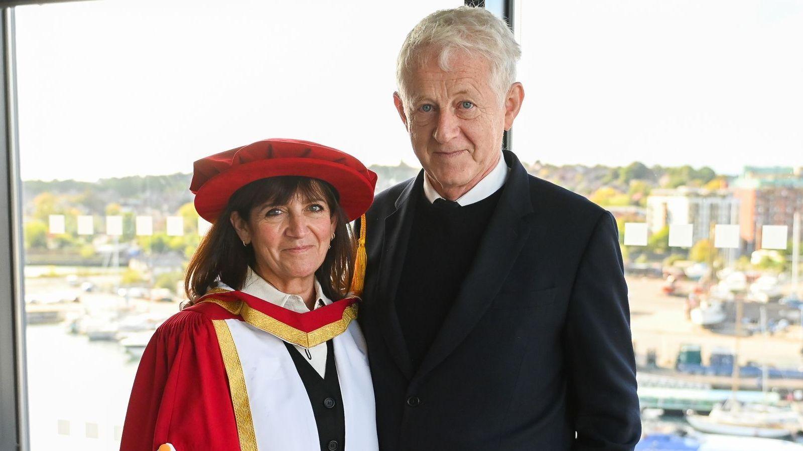 Emma Freud wears a red cap and a red, white and gold gown over a black jacket. She is smiling and stands next to her husband Richard, who wears a dark coloured jacket over a dark coloured jumper. They are both standing against a window and the scene behind is of Ipswich Marina, which is filled with boats.