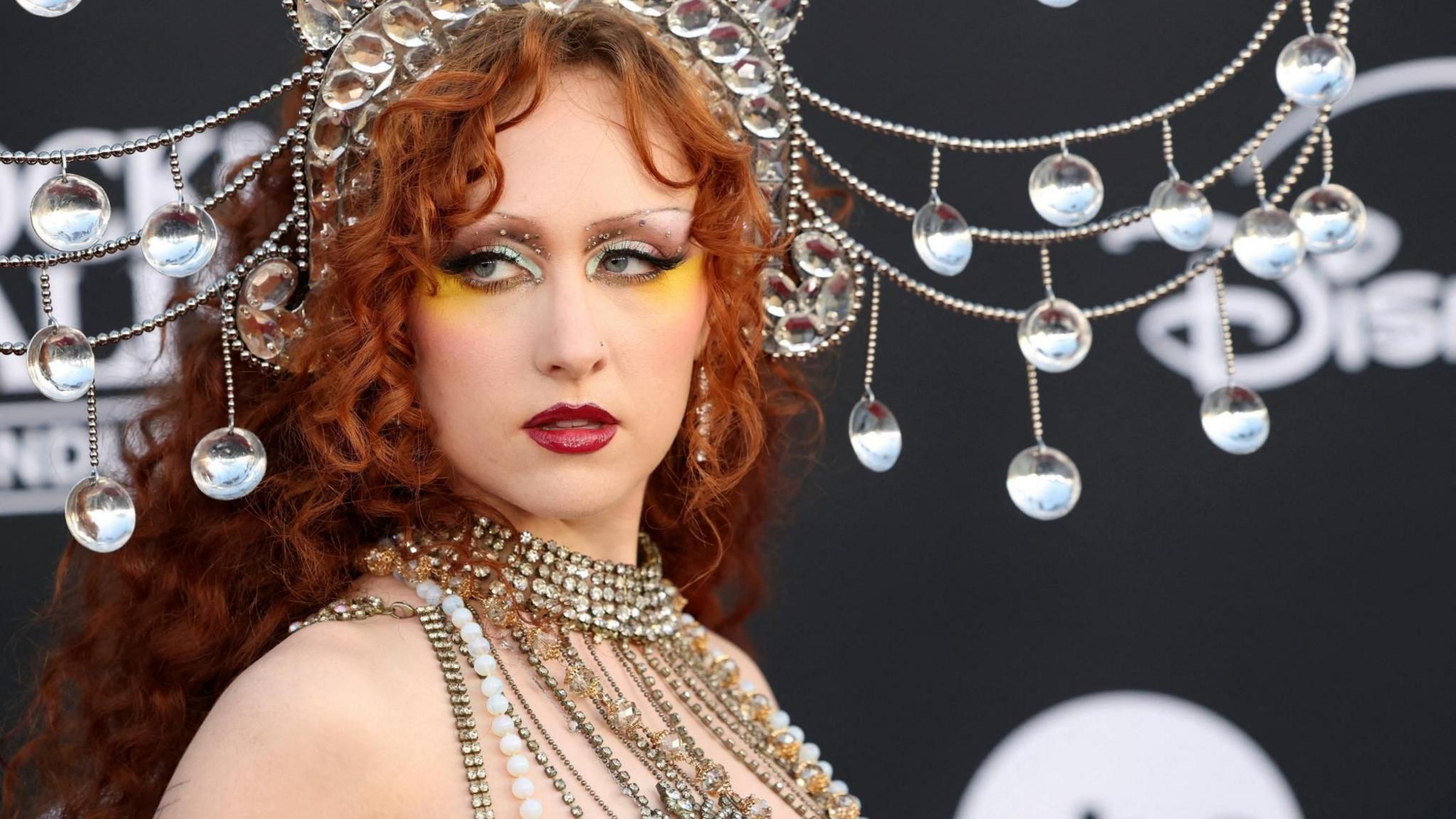 Close-up of a Chappell Roan wearing an ornate crystal headpiece and layered jeweled accessories, posing in front of a dark backdrop decorated with hanging reflective ornaments.