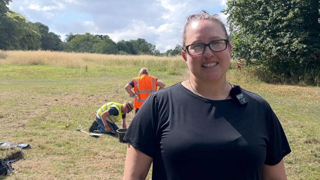 A woman with brown hair, tied back, and black-rimmed glasses smiles as she stands on a large common with green grass and trees. She is wearing a black T-shirt. Behind her, two people in high-vis vests are examining a piece of ground with a bucket standing between them.