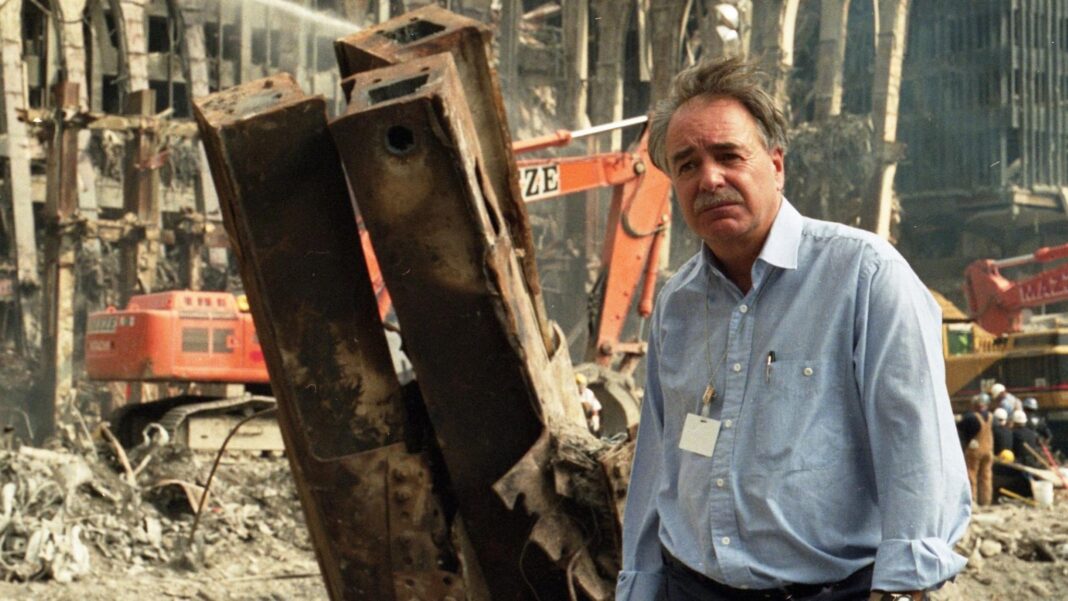 A man wearing a blue shirt and white ID badge stands in the destruction of the twin towers attack. There are metal girders sticking up and a digger behind them too.