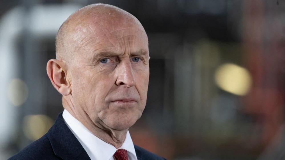 John Healey looks seriously at the camera with the background of the image out of focus. Healey is seen from the shoulders up and is dressed in a blue suit, white shirt and red patterned tie.