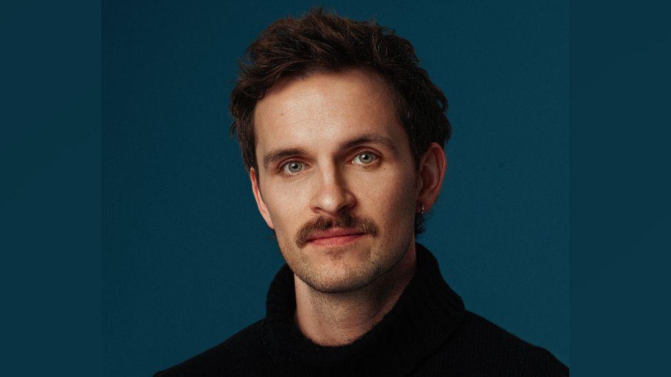 Vittorio looking at the camera. He is wearing a black jumper. He has brown hair and green eyes. The background is a blue/green colour.