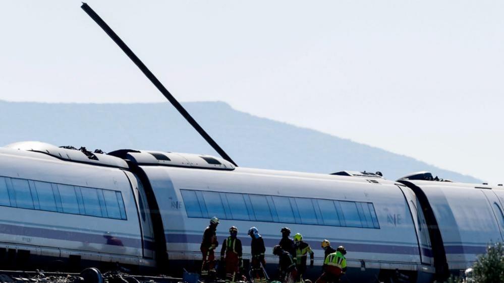 A piece of rail is seen emerging through the roof of a train carriage; there are emergency workers wearing high-visibility jackets and helmets in front of that carriage - one of three visible in the shot.