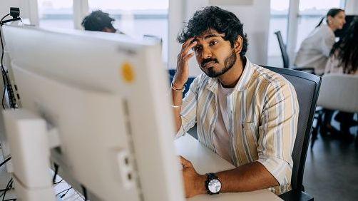 Representative image of a tired man working at office on desktop PC. His colleagues, most likely of different nationalities, are seen in a blurred background.