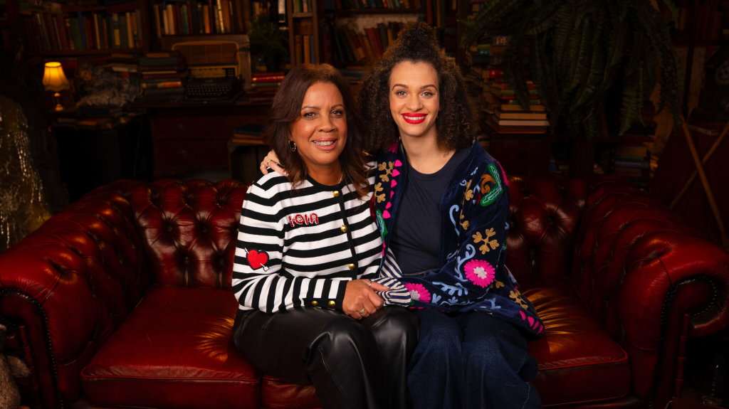 A woman wearing a black and white striped top, and a younger woman sitting next to her, are on a sofa, both smiling at the camera.