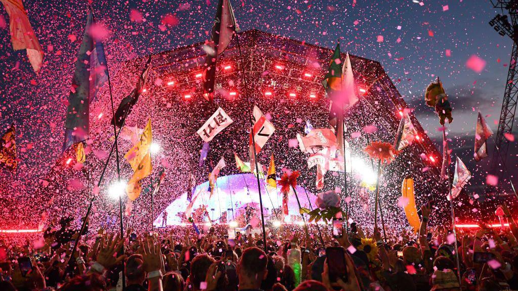 The Pyramid stage at Glastonbury festival is pictured lit up with a pink hue at night. There are thousands of pieces of confetti flying through the air as people in the crowd wave their arms and hold up tall flags.