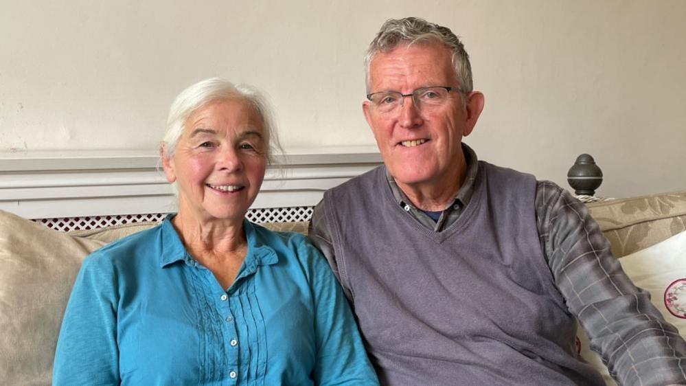 Bern Rogers and John Rogers are sat together smiling on their sofa. Bern is wearing a blue blouse, while John has glasses and is wearing a grey sweater vest and checked shirt.