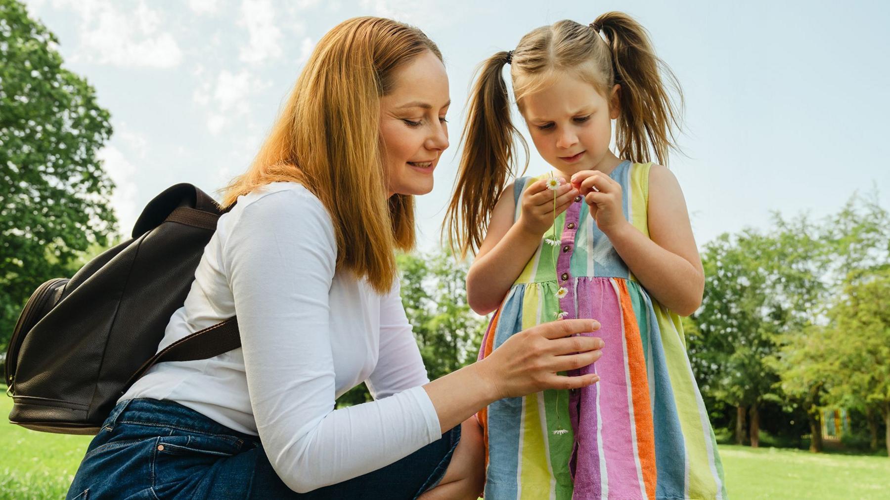 A little girl wearing a rainbow dress and looking down at a daisy chain in her hand. Her mother is crouching down next to her talking to her.
