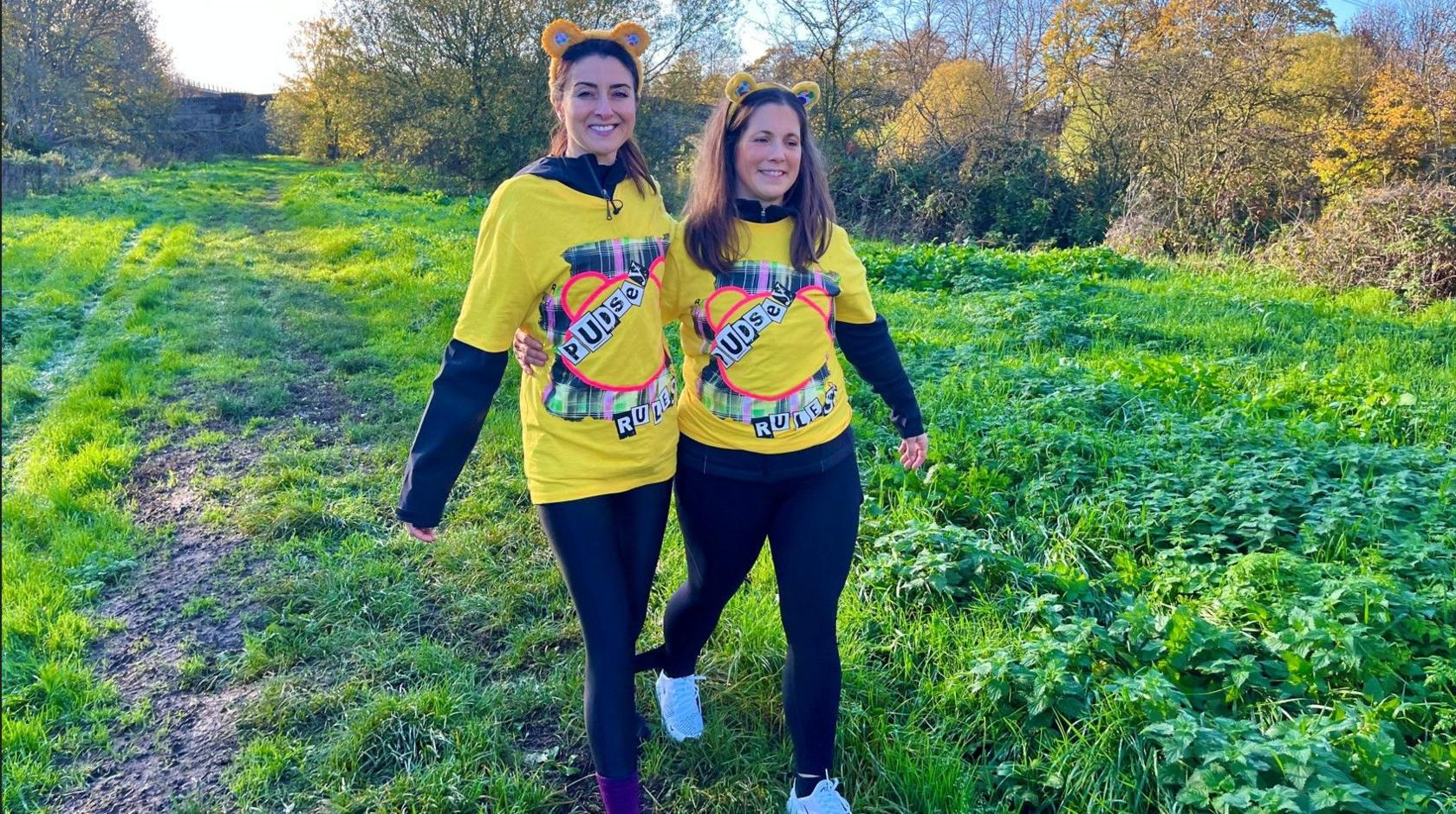 Amy Garcia (left) and Lindsey Burrow (right). Amy has black hair, a black top, a yellow Children in Need t-shirt and black leggings. Lindsey has brown hair, a black top, a yellow Children in Need t-shirt and black leggings. Both are wearing a yellow Pudsey ears headband. walking with their legs tied together for the Children in Need Three-Legged Challenge in 2025. They are walking through thick green grass with yellow autumnal bushes and trees either side.