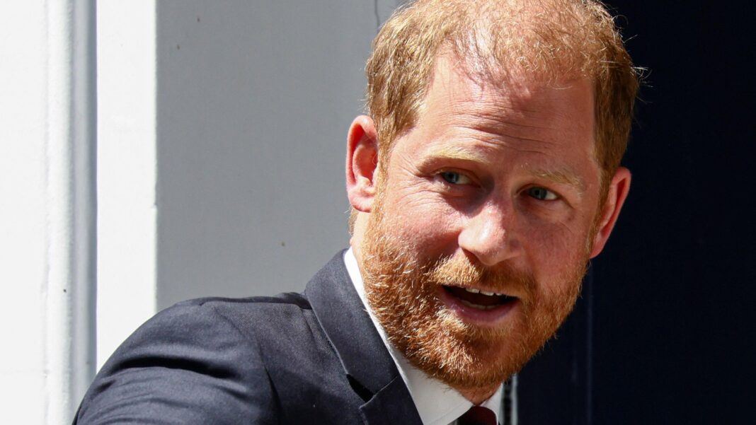 Prince Harry wears a black suit, white shirt and black tie as he walks through a white doorway.