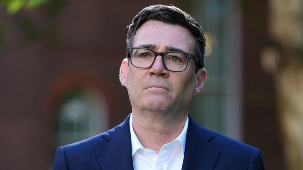 Andy Burnham looks in the dustance as he wears a navy blue suit and white shirt with a blurred background.