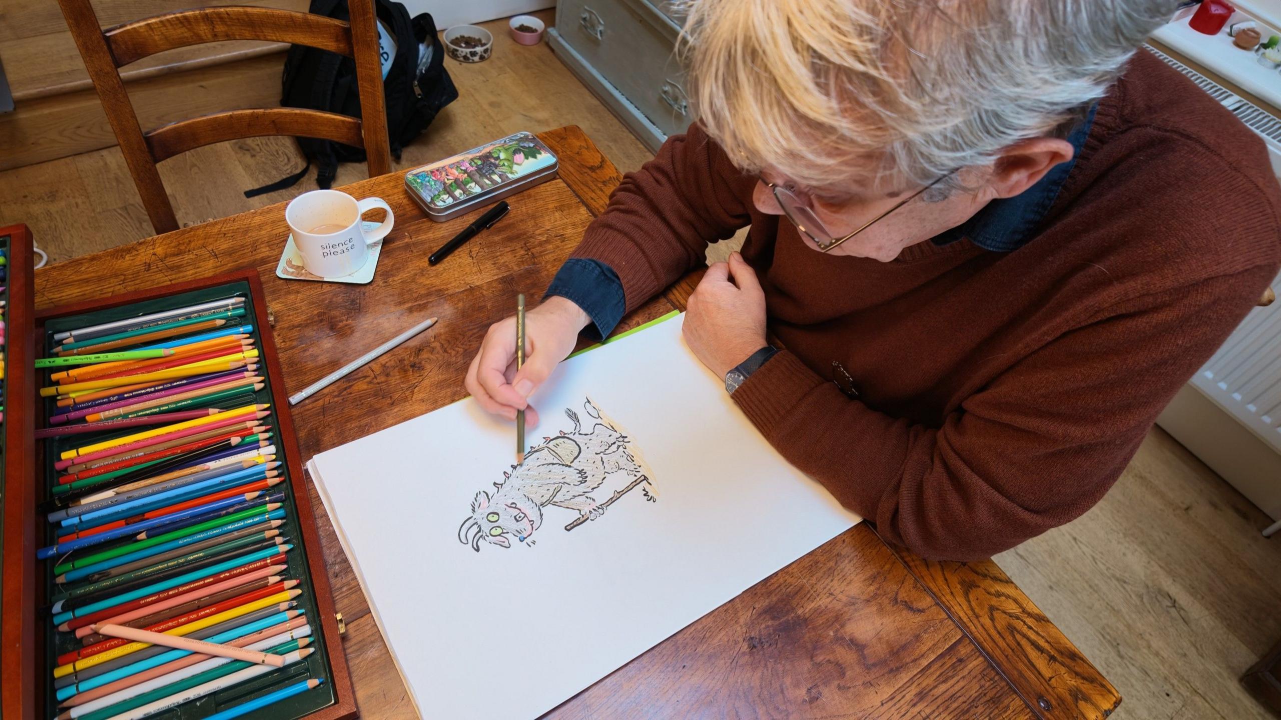 High-angle view of Axel Scheffler sitting at a wooden table with a large box of coloured pencils, hand-drawing a character on a white sheet of paper.