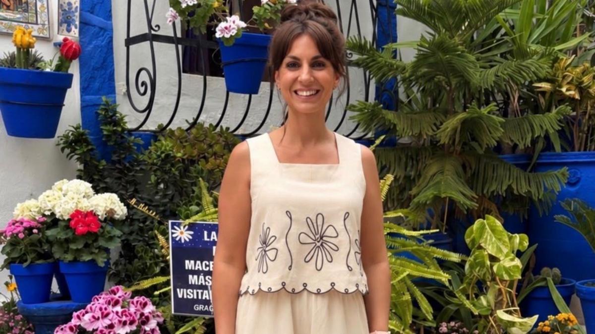 A woman with brown hair tied in a bun, wearing a cream-coloured matching linen set with dark floral embroidery stands in front of a wall with flowers in pots and leafy green plants behind her.