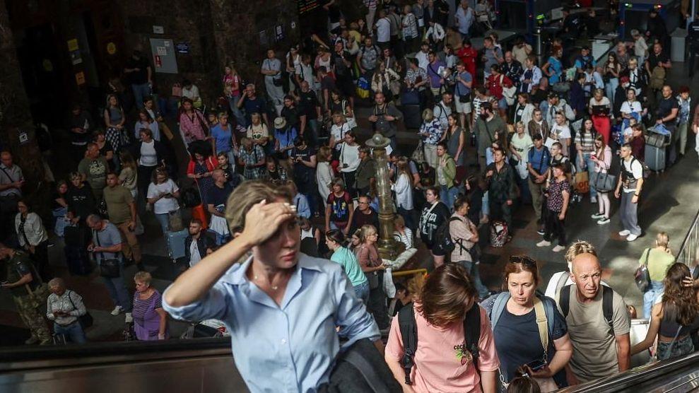 Several people stand on an escalator going up as dozens more passengers stand around in the main concourse of a train station in Kyiv