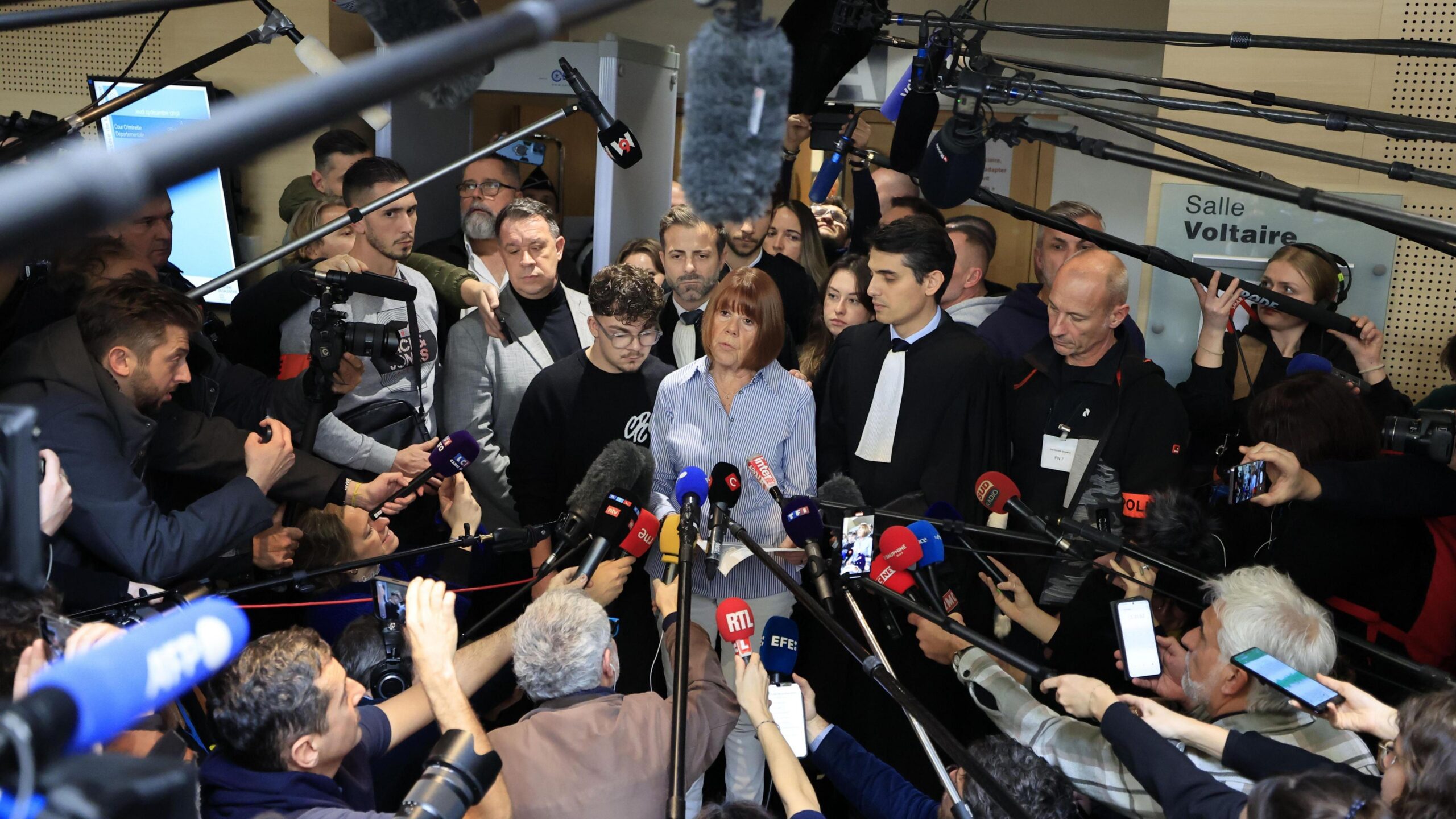 Gisèle Pelicot framed at the centre of a dramatic wide-angle shot inside the courthouse, with a huge crowd of journalists surrounding her with microphones