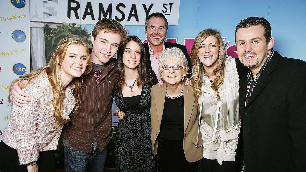 Seven people stand with their arms around each other's shoulders in front of a partition with the graphic of a street sign saying "Ramsay St"