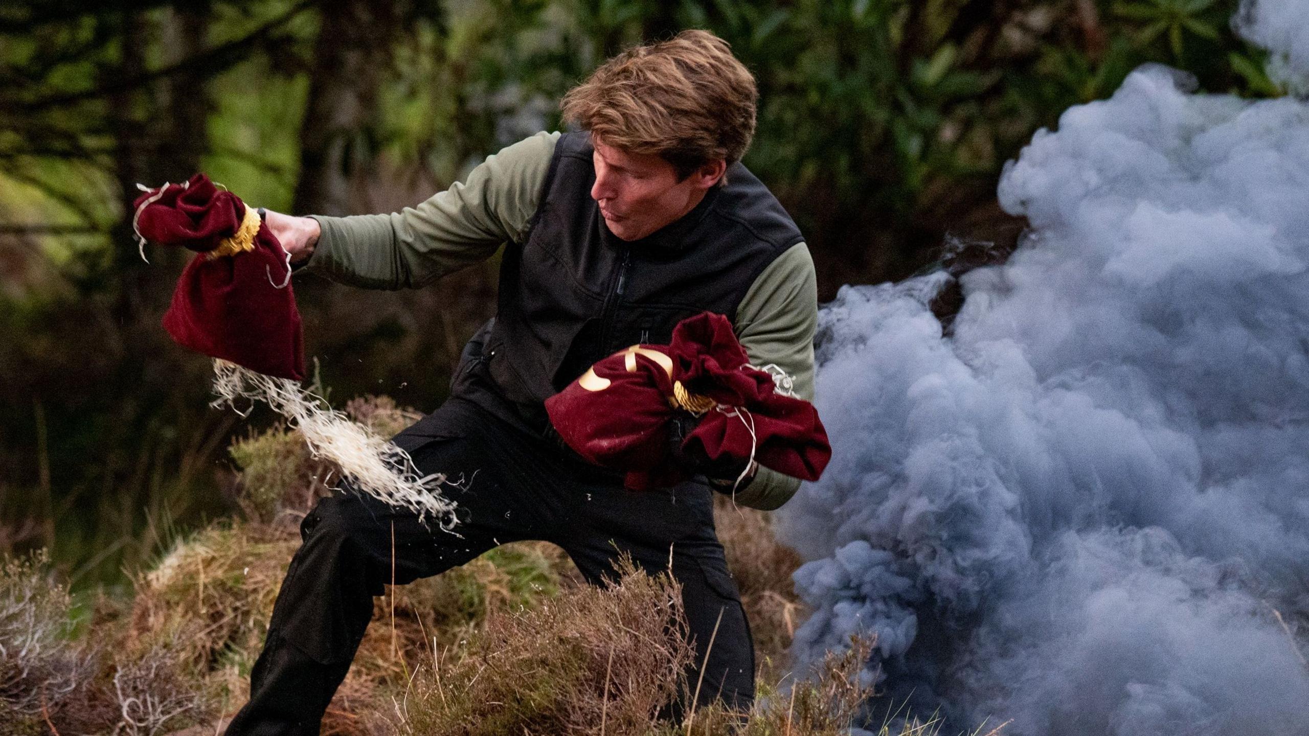 James Blunt pulling two red money bags out of some grey smoke in some woods