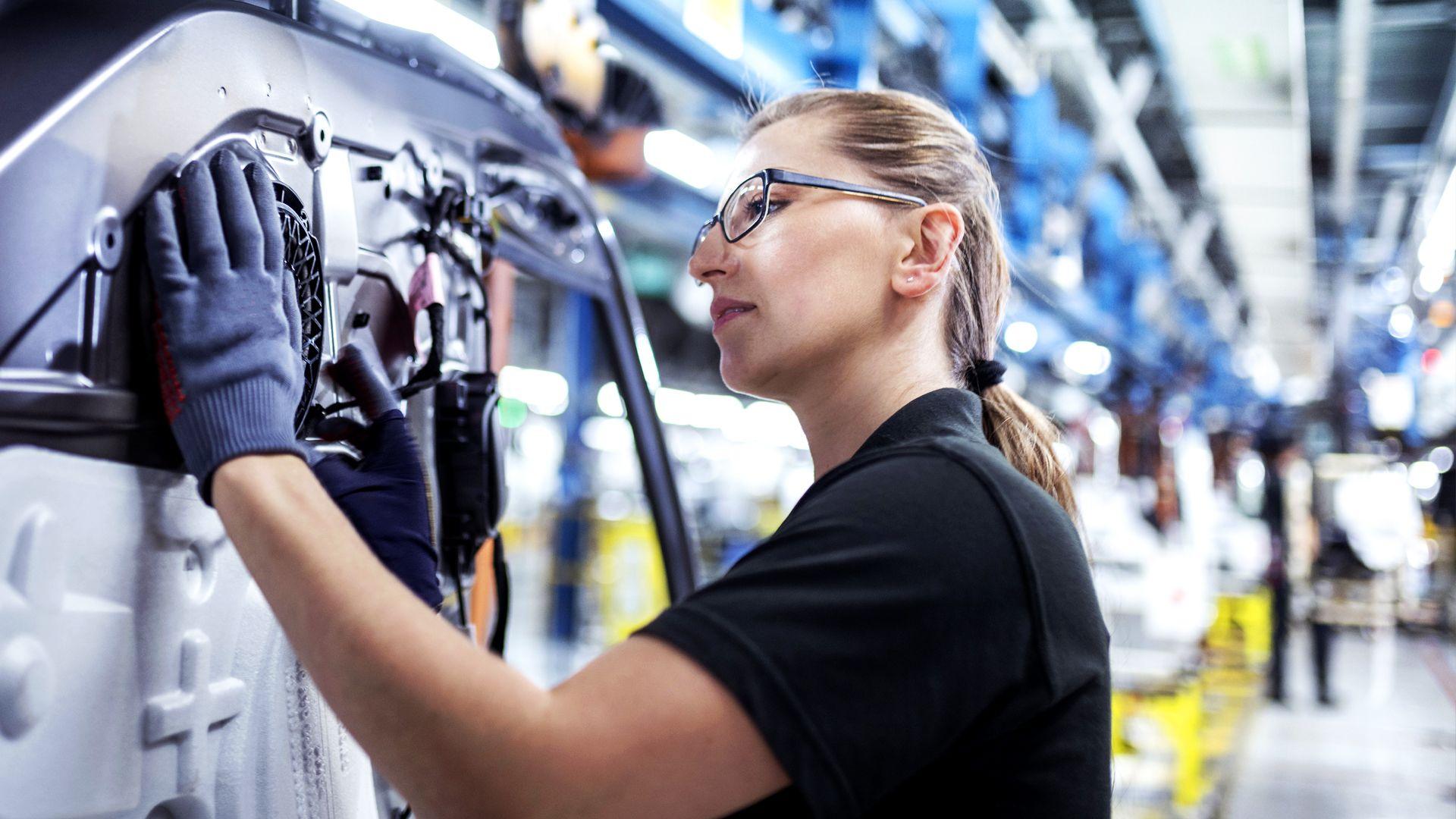 A woman in a black T-shirt and wearing glasses and protective gloves works on a machine in a factory