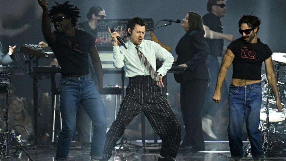 A man with short brown hair and wearing pinstripe trousers and a shirt and tie is dancing on stage, with two dancers to either side of him and three musicians behind.