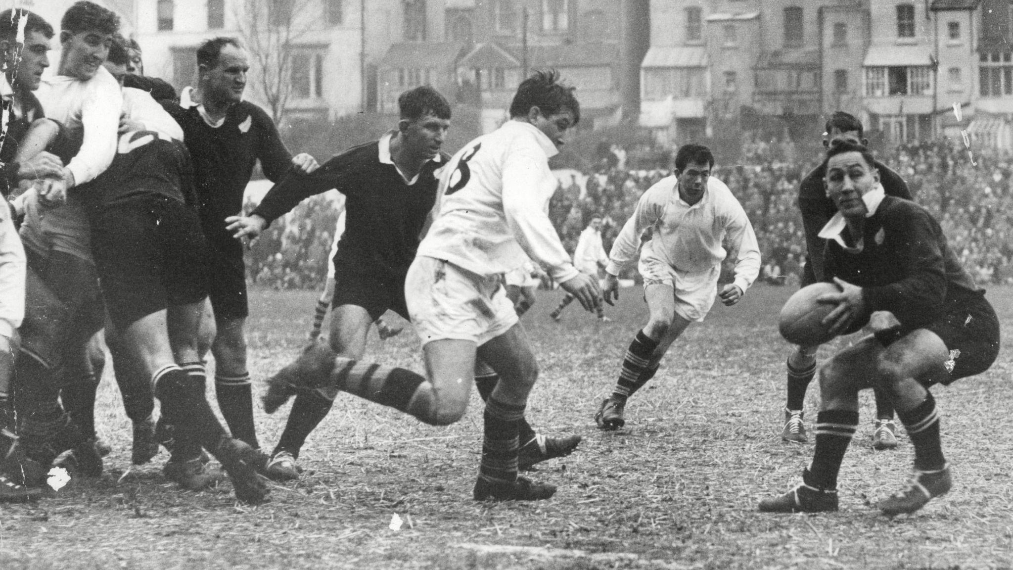 New Zealand players in black and Swansea players in white playing on a muddy pitch in front of a packed crowd