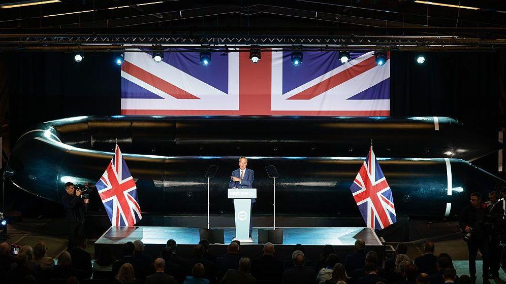 Nigel Farage stands at a podium in front of a large black inflatable boat, with Union flags in the backdrop and either side of him and light blue lighting.