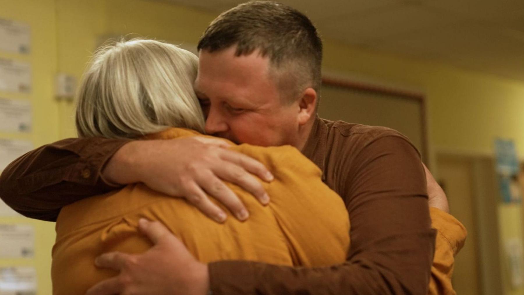 A white man with short brown hair wearing a brown shirt hugs a woman with short blonde hair wearing a dark yellow shirt in a corridor with yellow walls