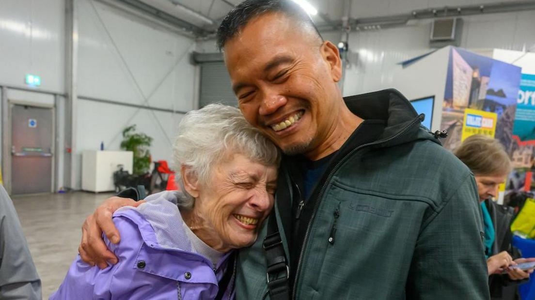 A woman with grey short hair in a purple raincoat is hugging a man with short brown hair in a green hoody. They are both smiling with their eyes closed.