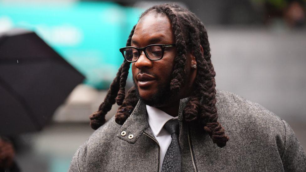 Omololu Akinlolu arriving at Southwark Crown Court, south London on Monday October 26, 2026. He has shoulder-length hair, glasses and is wearing a grey herringbone jacket, white shirt and grey tie.