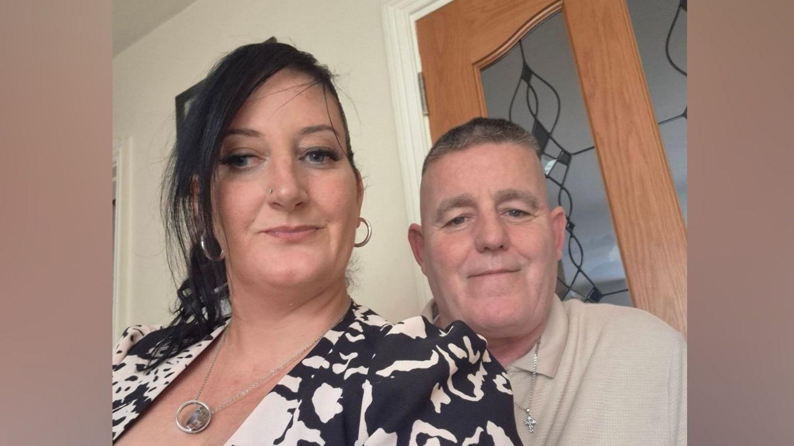 Kim English - a woman with long, black hair swept back from her face - looks to the camera in a selfie-style photo.  Her husband Philip English is standing close to her smiling.  He has short grey hair and is wearing a beige polo shirt and a silver necklace with a cross-shaped pendant.  She is wearing a black and white patterned jacket and a silver necklace.
