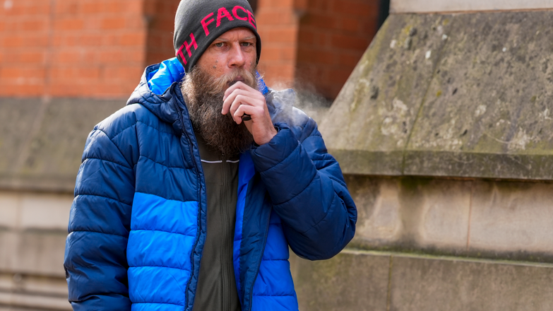 A man in his 30s, with a large beard, grey woollen hat and blue bubble coat, stands outside a stone building holding a black vape to his mouth.