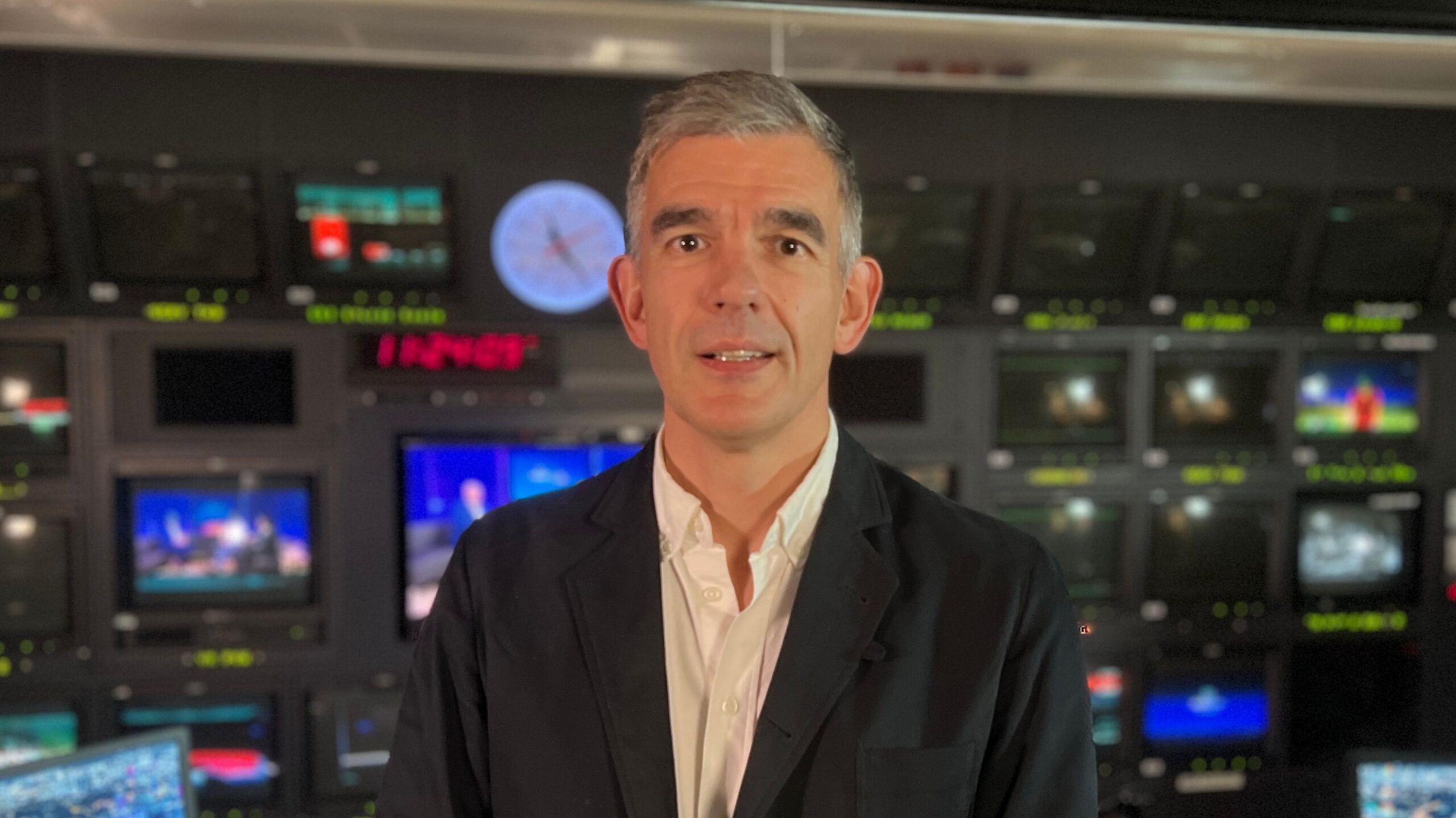 A photo of a man wearing a black suit jacket with a white shirt looking into the camera. He is in a TV gallery and has a half smile. He has grey hair.