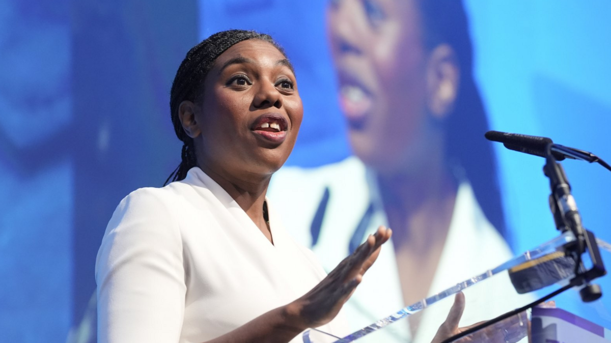 Head and shoulders shot of Badenoch speaking a a glass podium. She is wearing a white top and has her black hair tied back in a low ponytail.