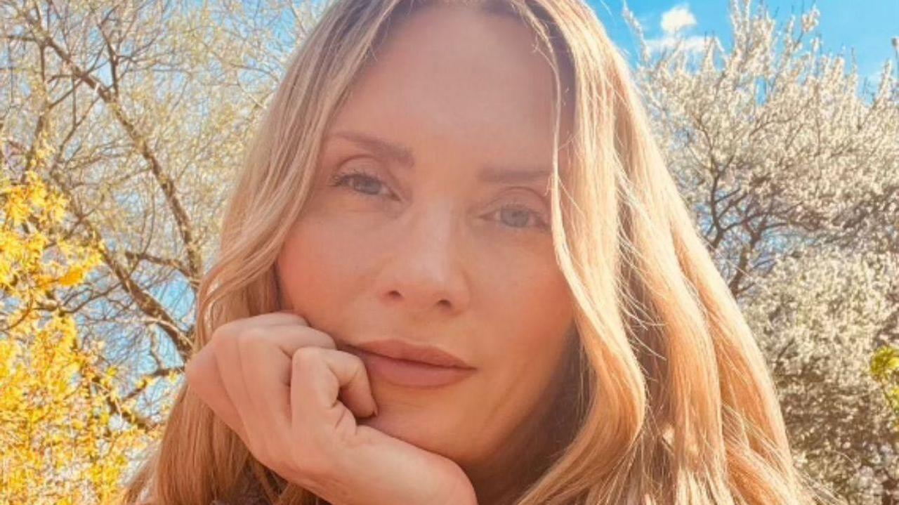 Bernadette Spofforth looks into the camera with her hand under her chin. She has wavy blonde hair.