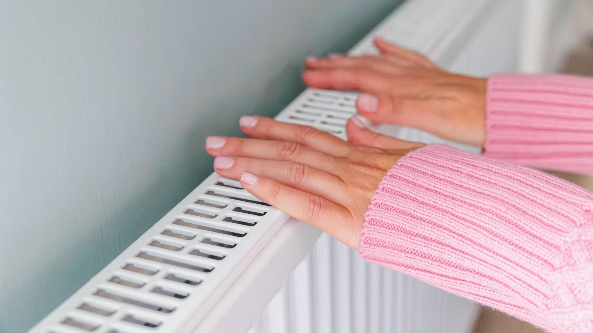 A woman touching the top of a radiator with two hands. She is wearing a long-sleeved pink knitted jumper and has neatly manicured nails.