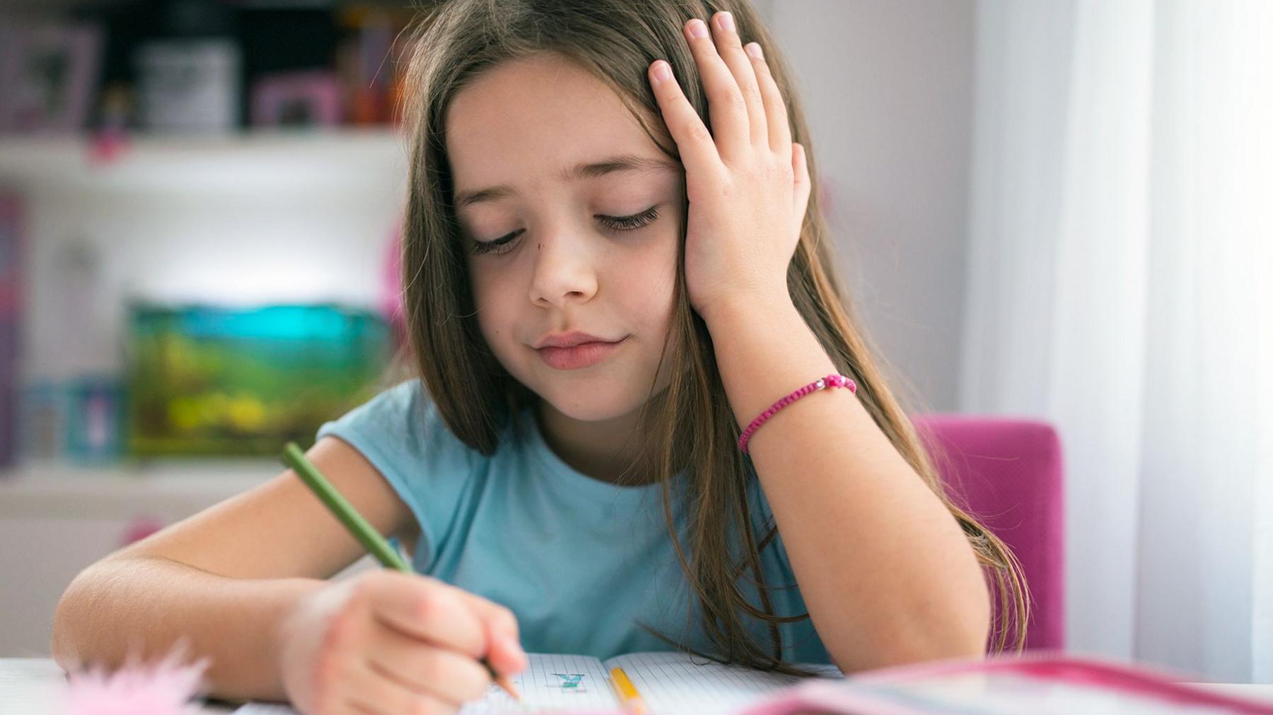 A little girl aged five to seven wearing a light blue T-shirt, sitting at a desk drawing with a green pencil and resting her head on one hand