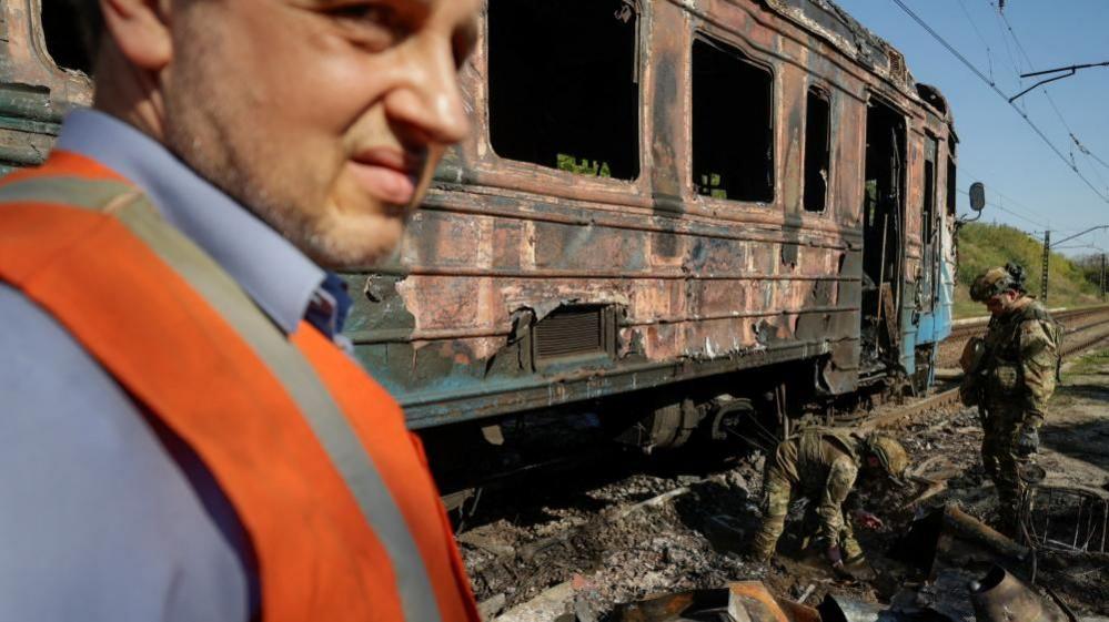 A man wearing a blue shirt and orange high vis jacket stands in front of a burnt train carriage