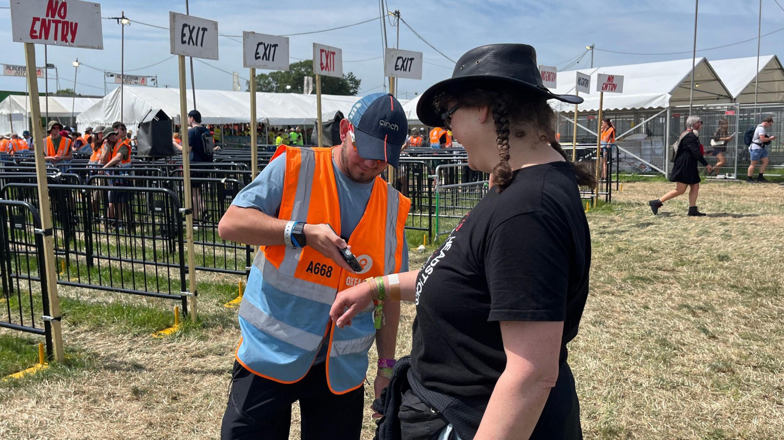 A man in hi-vis stamps woman's hand in a field in front of festival gate. There are different lines fenced off behind him with signs for no entry and exit.